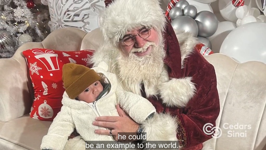 Santa Claus holding a baby dressed in a cream-colored outfit and brown hat.