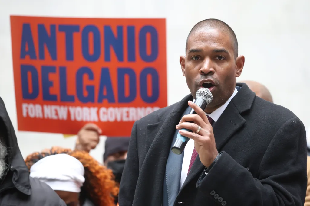 Lt. Governor Antonio Delgado speaking into a microphone with a red campaign sign behind him.