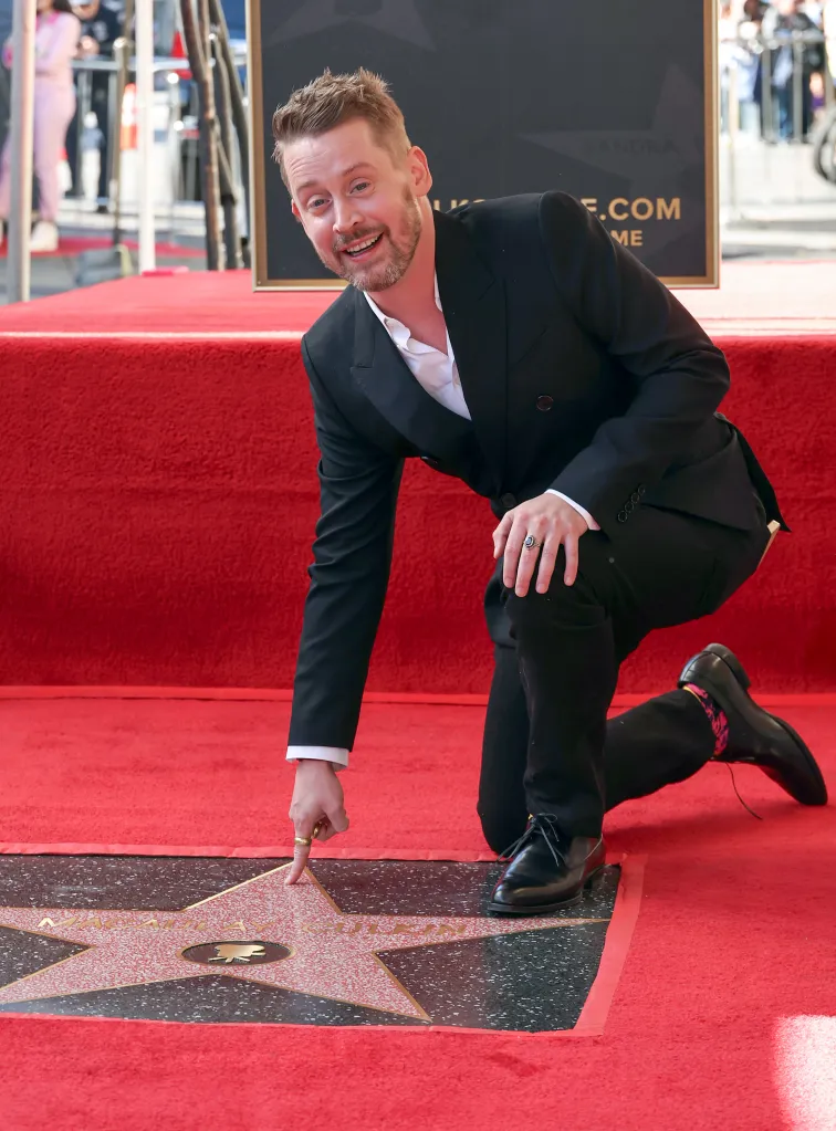 Macaulay Culkin kneeling on a red carpet, pointing to his newly unveiled star on the Hollywood Walk of Fame.