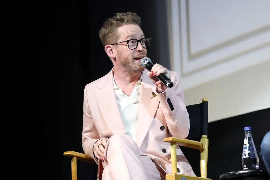 Macaulay Culkin speaking onstage in a pink suit, holding a microphone.