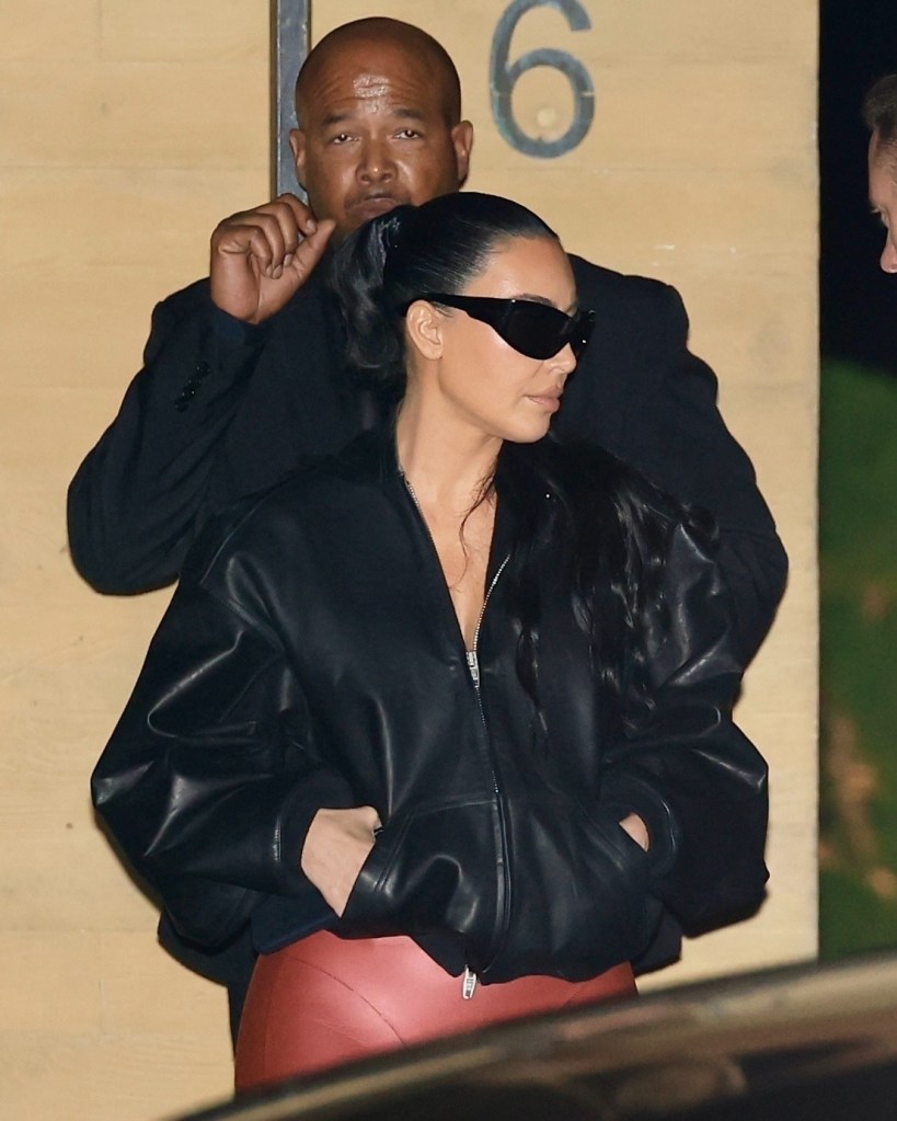 Kim Kardashian in a black leather bomber jacket and red pants with a man behind her.