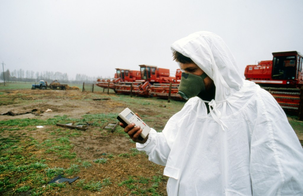 Man in a white protective suit and gas mask measuring radioactivity with a Geiger counter near red machinery in Chernobyl.