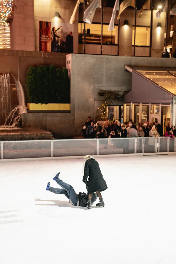 A man has fallen on his back on an ice skating rink while a woman stands over him during proposal at Rockefeller Center