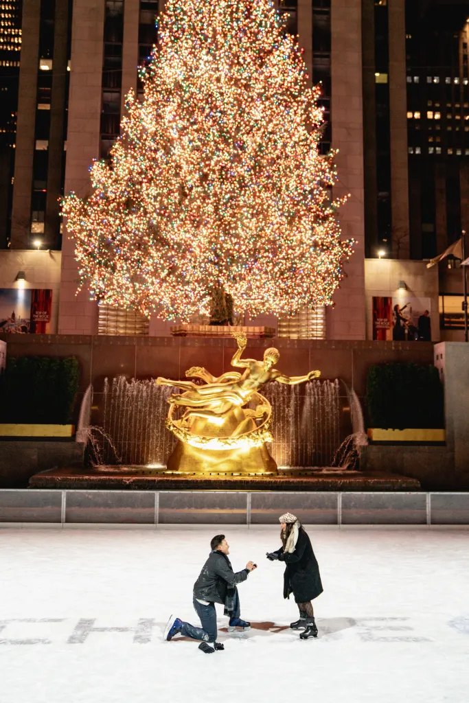 A man proposes to a woman on an ice rink at night, with a large illuminated Christmas tree and golden statue in the background.