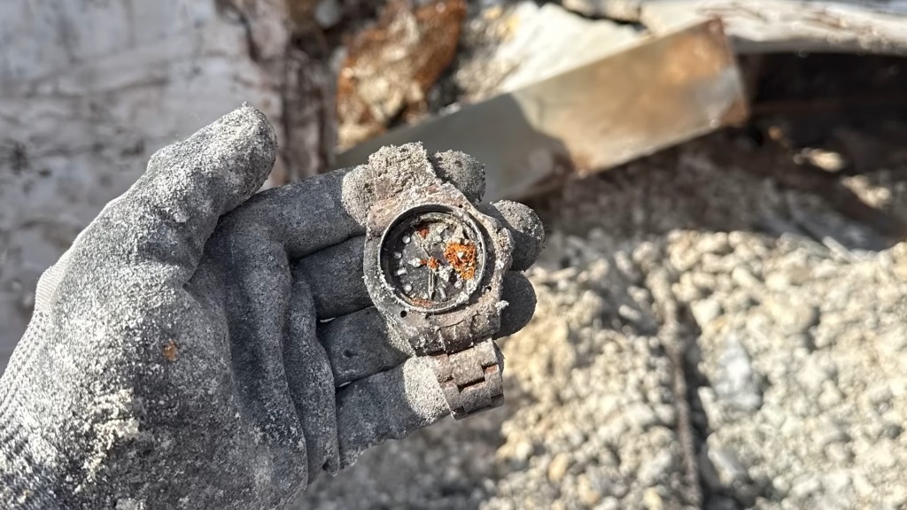 A gloved hand covered in debris holds a fire-damaged Rolex Deepsea watch.