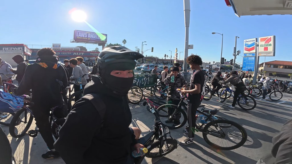 A person in a black balaclava and helmet looks at the camera in a crowd of people with bicycles at a Chevron gas station.