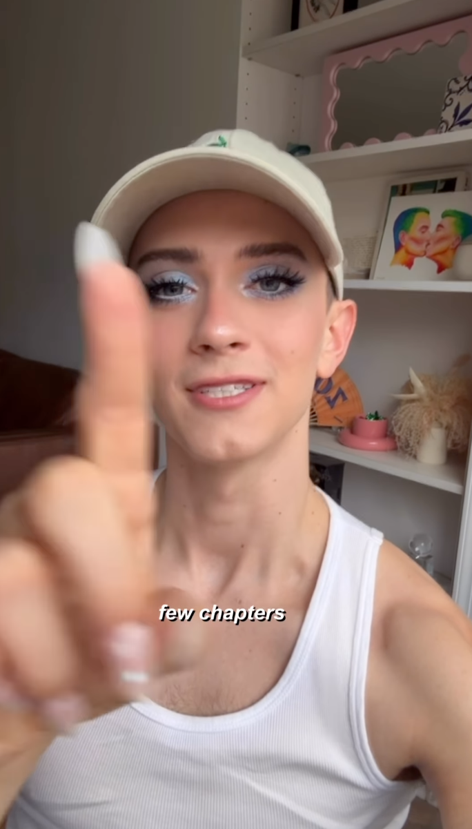 Matt Bernstein in a white tank top and baseball cap, wearing blue eyeshadow, holding up a finger to the camera.