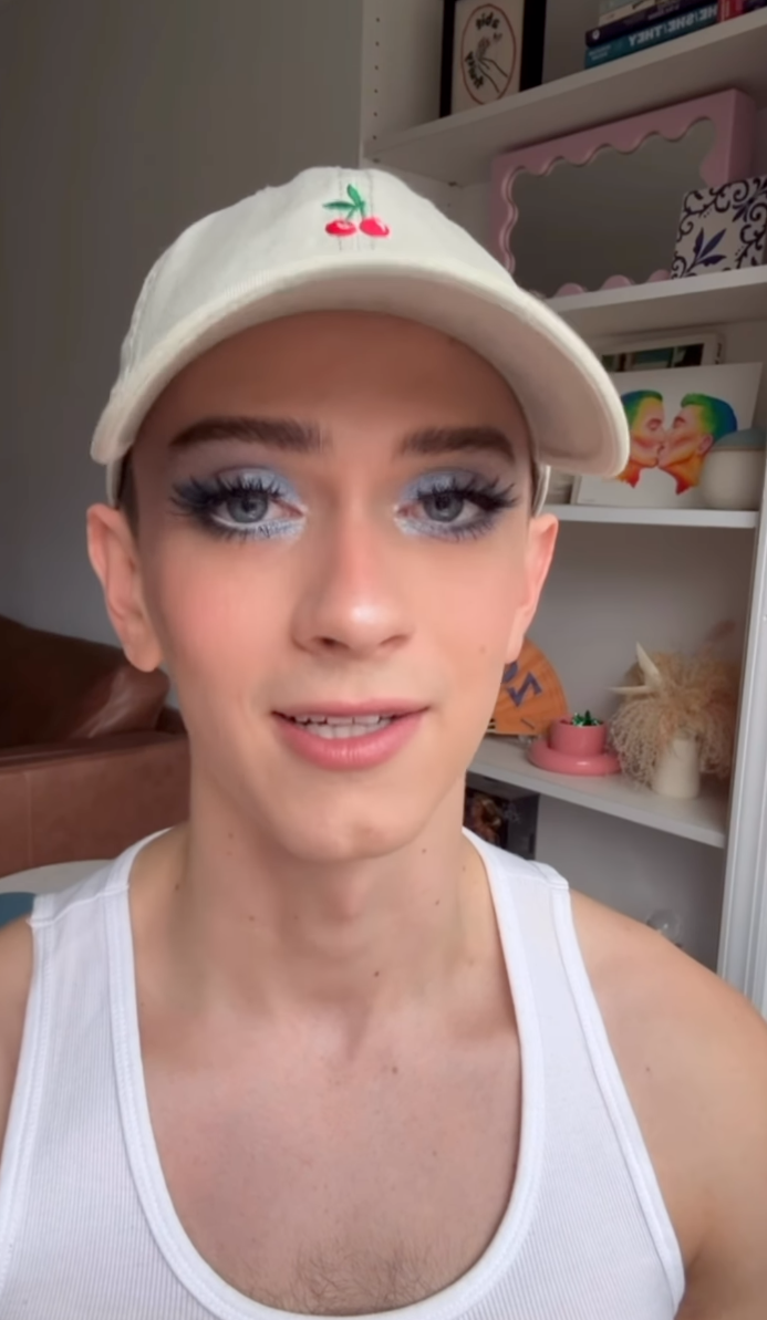 Matt Bernstein wearing blue eye makeup and a white cap with two cherries.