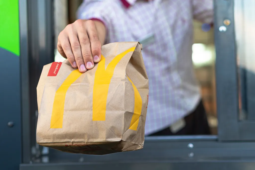 A McDonald's worker's hand holding a paper fast food bag through a drive-thru window.