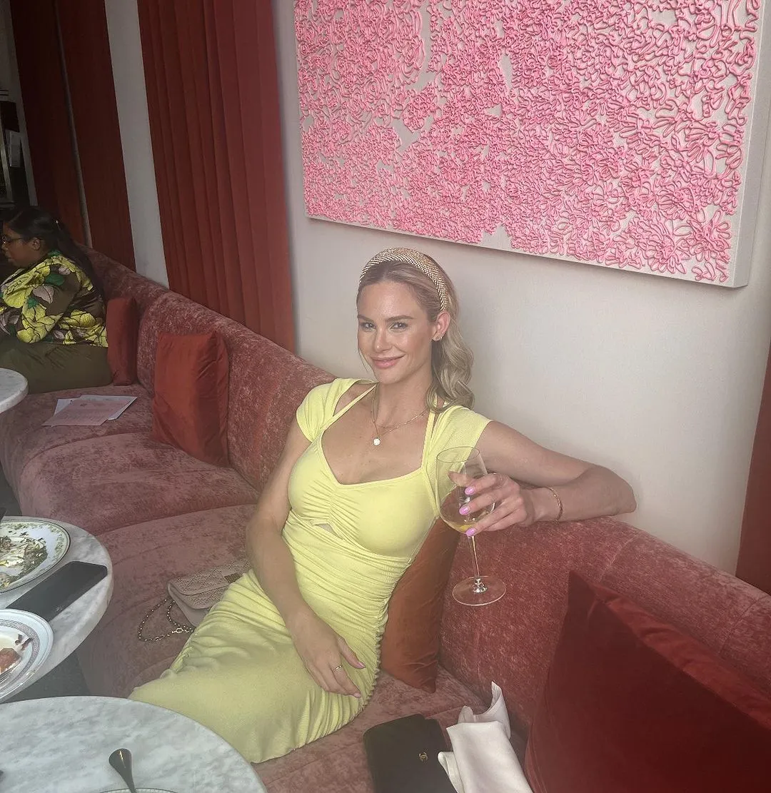Meghan King in a yellow dress, holding a glass of white wine, seated on a red sofa.