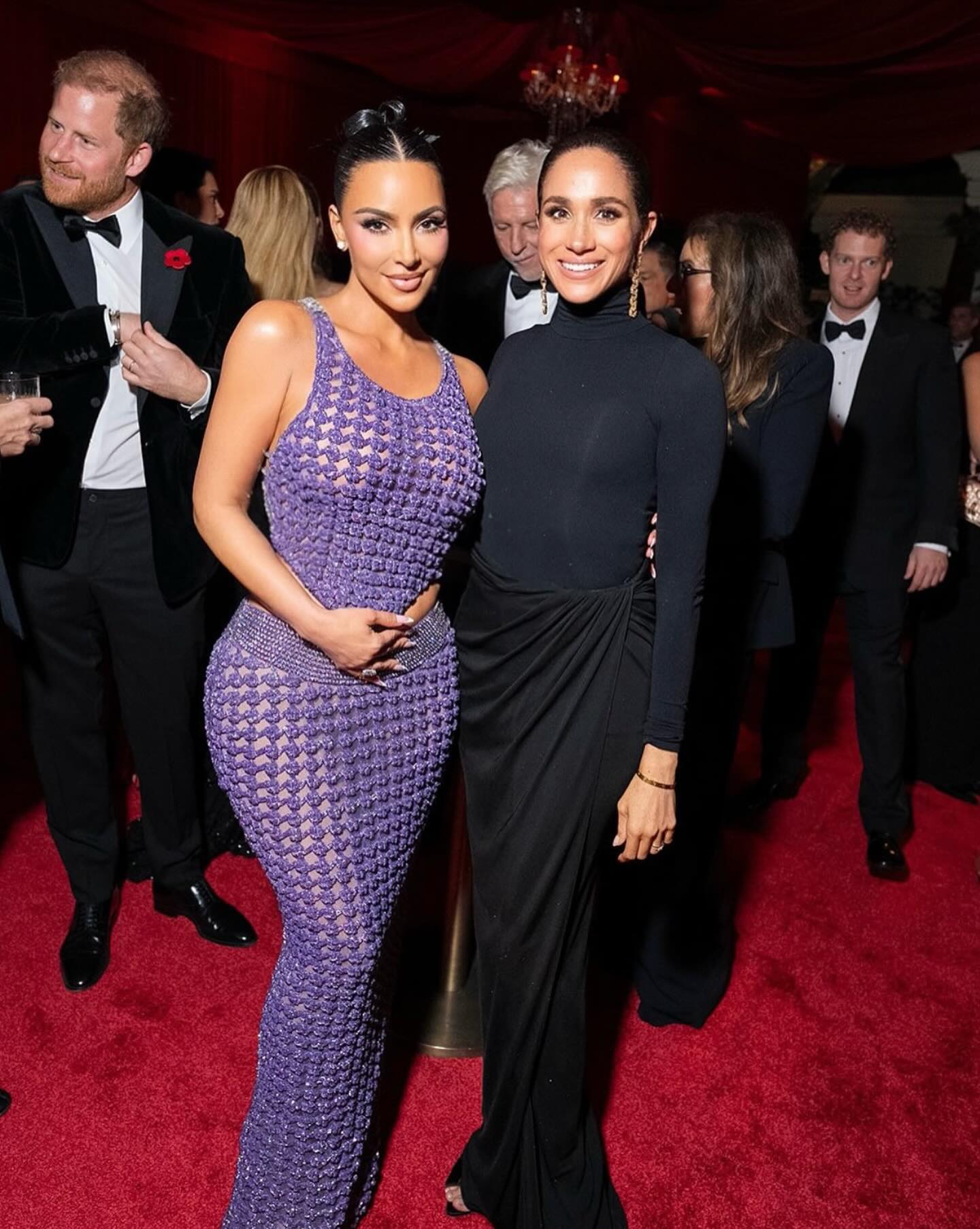 Meghan Markle and Kim Kardashian posing together on a red carpet with Prince Harry in the background.