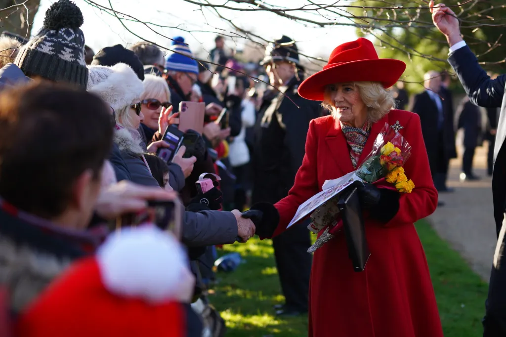 Queen Camilla in a red coat and hat shaking hands with a member of the public.