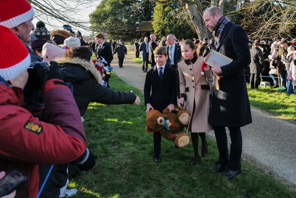 Prince William, Prince Louis, and Princess Charlotte walking among crowds of people, with Prince Louis holding a large teddy bear.