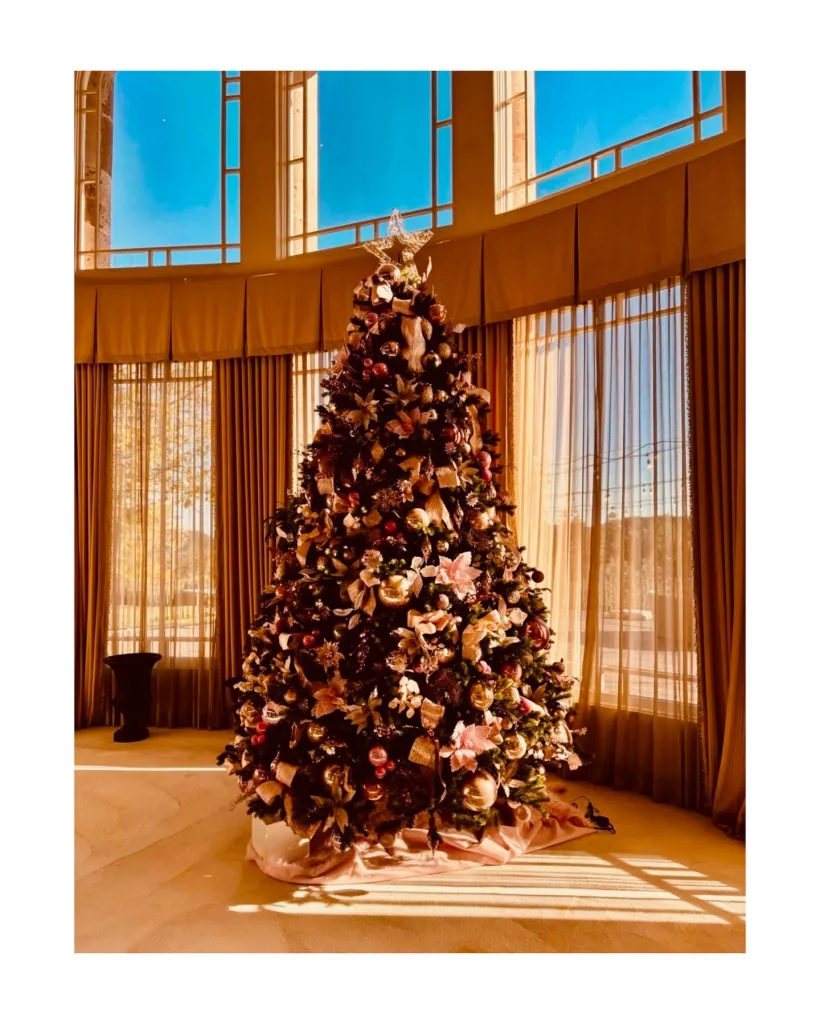 A Christmas tree adorned with gold, pink, and red ornaments, ribbons, and a star topper, standing indoors by a large curved window with gold curtains.