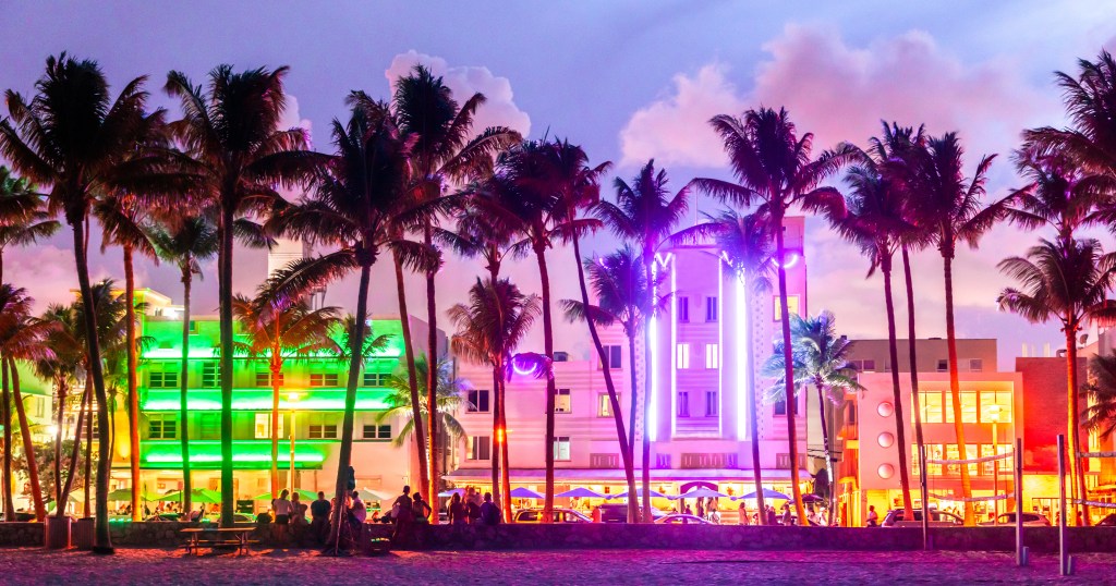 Art Deco buildings on Ocean Drive in Miami Beach, illuminated at night with neon lights, surrounded by palm trees.