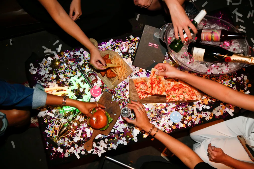 A party table from an overhead view with hands reaching for pizza, a burger, and chicken strips with fries, surrounded by colorful confetti, disco balls, and a bucket of champagne.