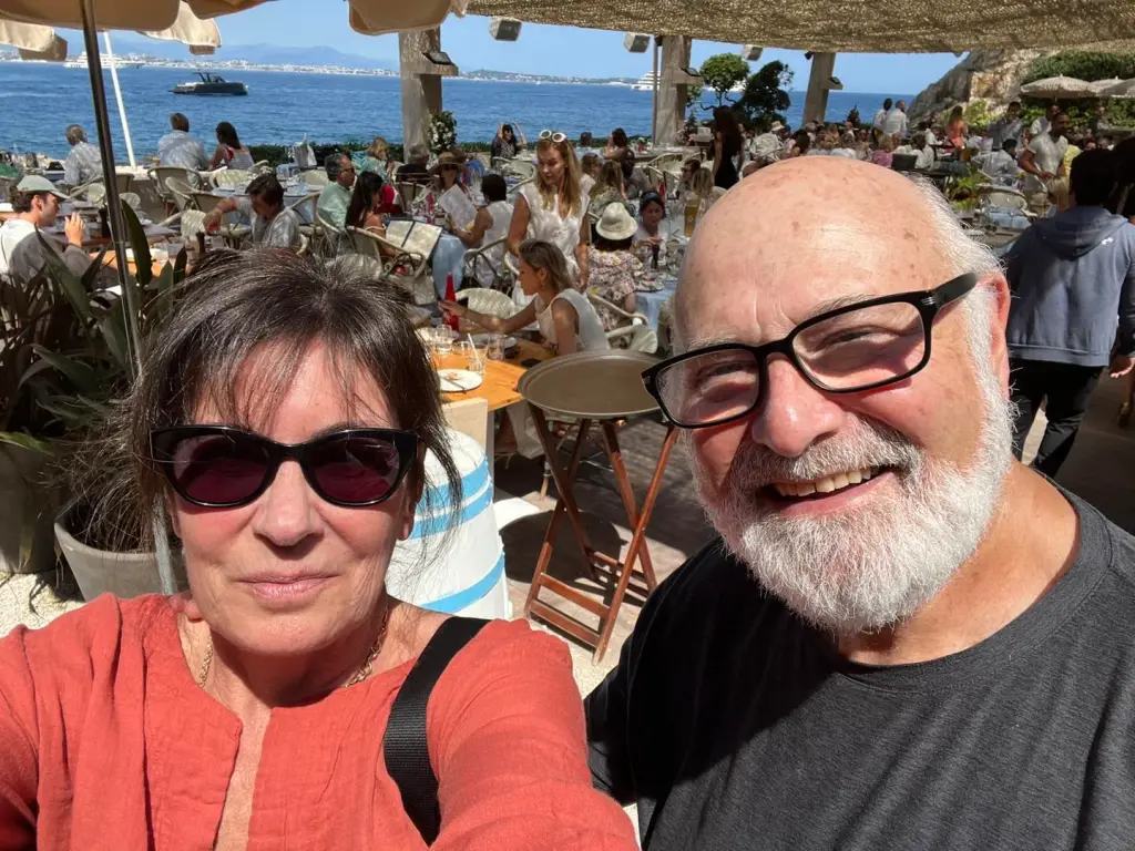 Michelle and Rob Reiner smiling at the camera with a busy restaurant by the ocean in the background.
