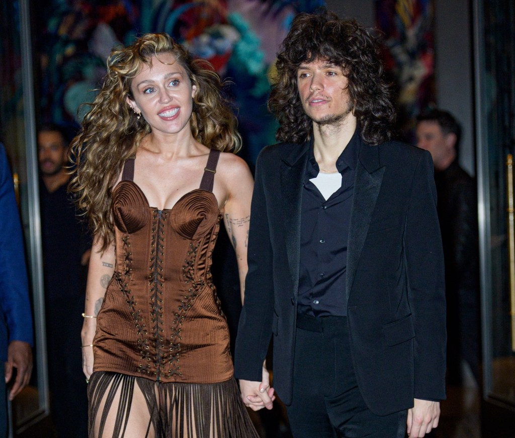 Miley Cyrus in a brown fringe dress and Maxx Morando in a black suit, holding hands and smiling.
