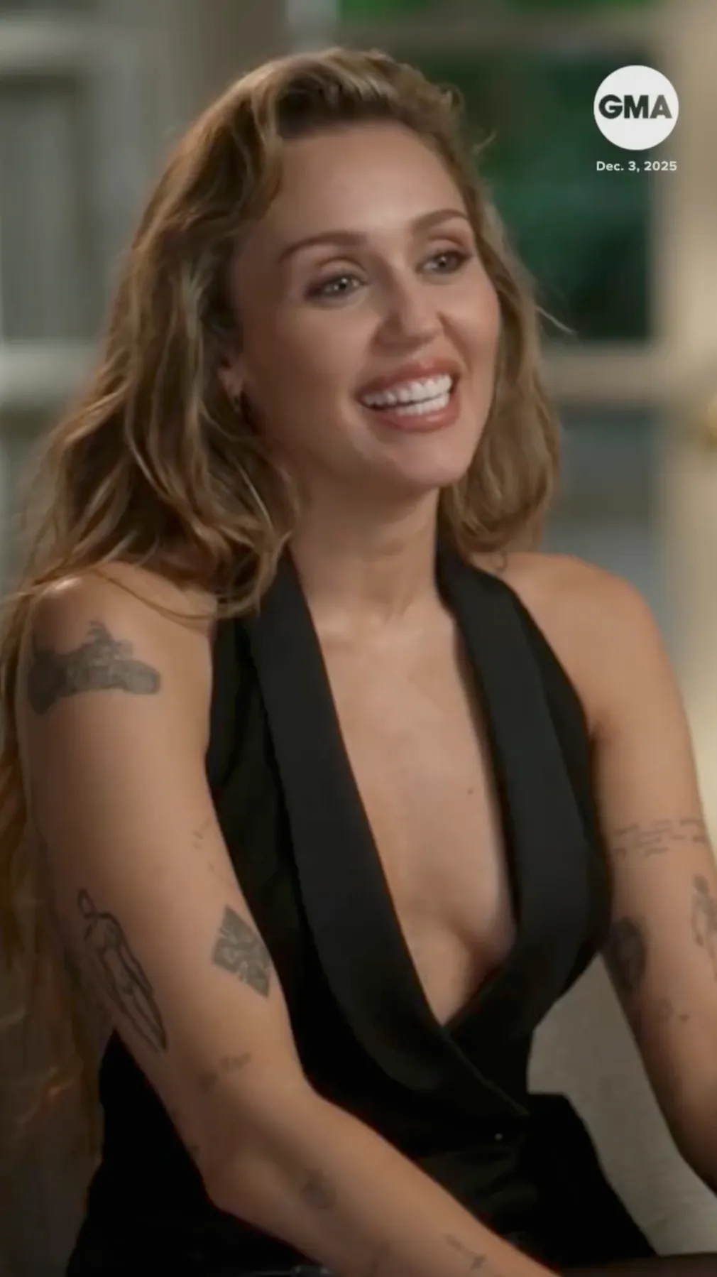 Miley Cyrus smiles while wearing a black top and showing off her arm tattoos in an interview with 