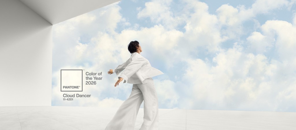 A person in white clothing standing on a white surface with a blue sky and white clouds in the background, next to a Pantone Color of the Year 2026 