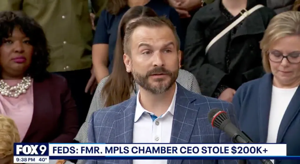 A man with a beard in a blue plaid suit jacket speaking into a microphone, flanked by two women, with a news chyron at the bottom reporting