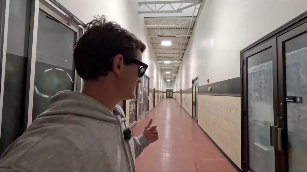 A person in a gray hoodie and sunglasses stands in a long, empty hallway with glass-fronted rooms on either side.