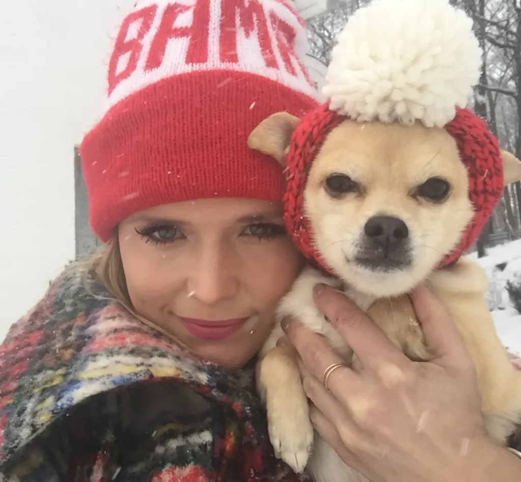 Miranda Lambert holding her dog, both wearing red hats, in falling snow.