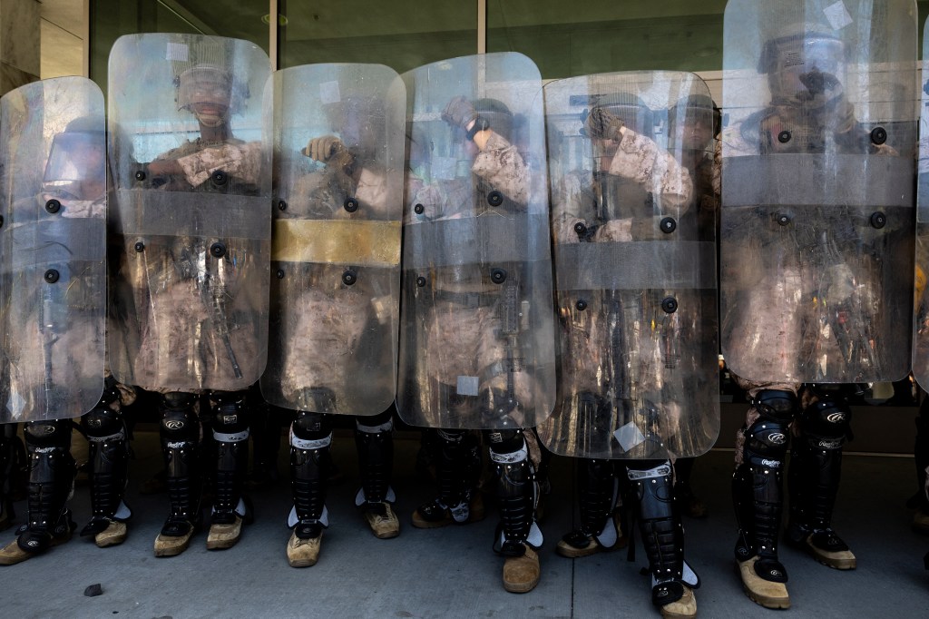 US Marines and National Guardsmen in camouflage uniforms with riot shields and leg protection stand at the entrance of a building.
