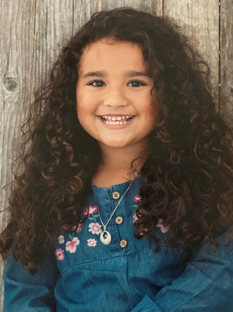 A smiling Melodee Buzzard, a young girl with long, curly dark hair, wearing a denim dress with embroidered flowers and a necklace with a round pendant.