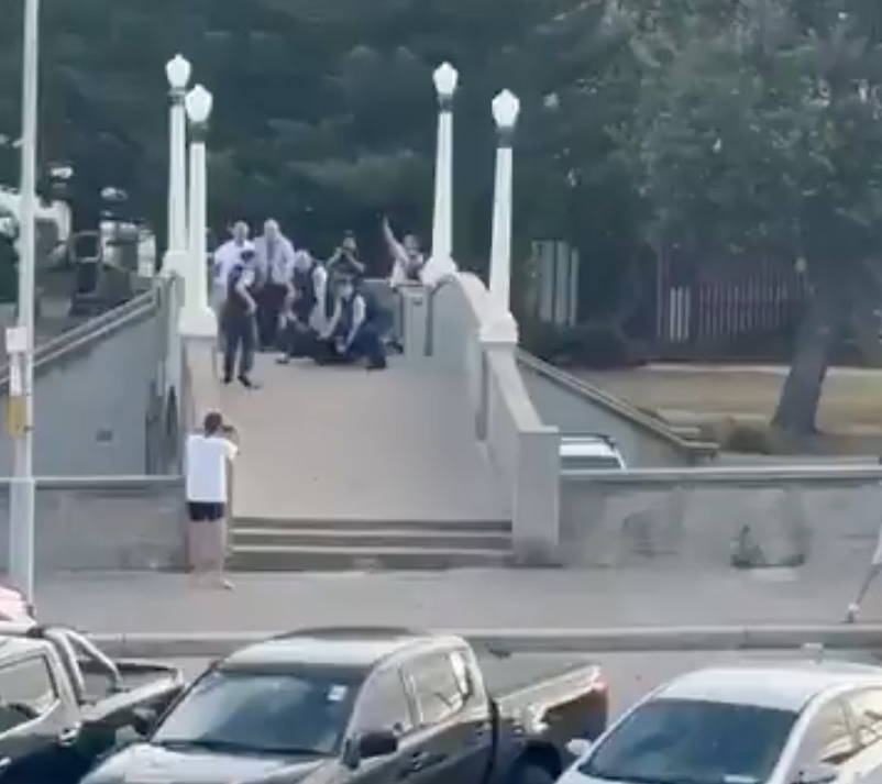 A man on a pedestrian bridge with his hands raised, surrounded by police and bystanders, after confronting an alleged shooter.