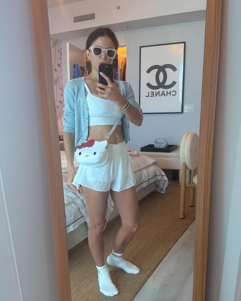 Bethenny Frankel taking a selfie in a mirror, wearing white athletic wear, a blue cardigan, and a white Hello Kitty purse.