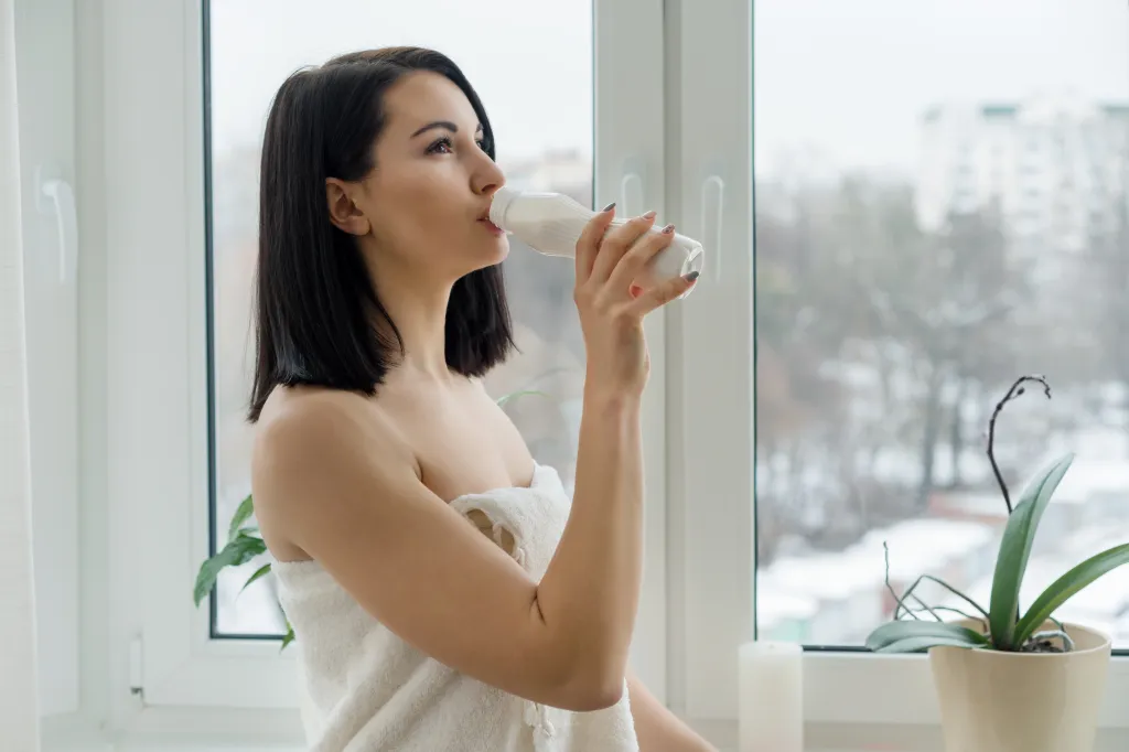 Young woman in a bath towel drinking milk or yogurt from a bottle by a window.