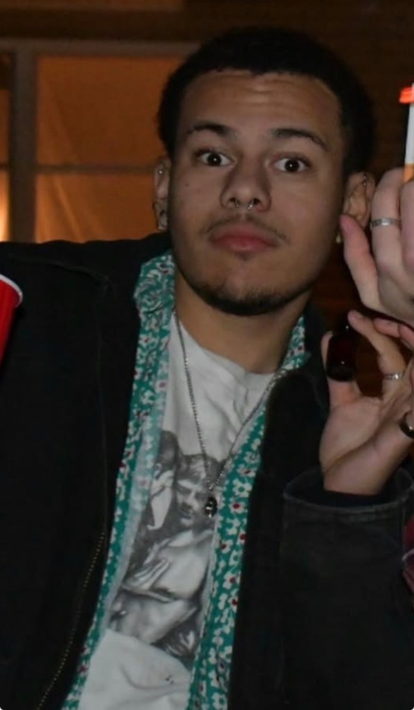 A young man with dark, curly hair, a light mustache, and a soul patch, wearing a septum piercing, looking at the camera with wide eyes and open mouth, wearing a floral collared shirt with a T-shirt underneath.