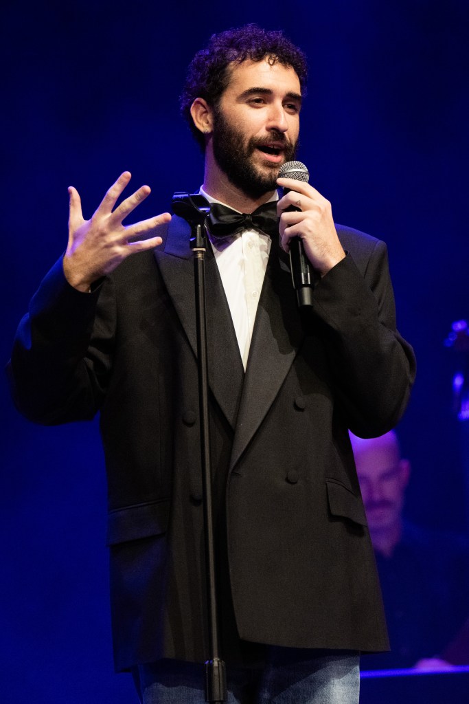 Musician Jordan Firstman performing onstage, holding a microphone in one hand and gesturing with the other, wearing a tuxedo.