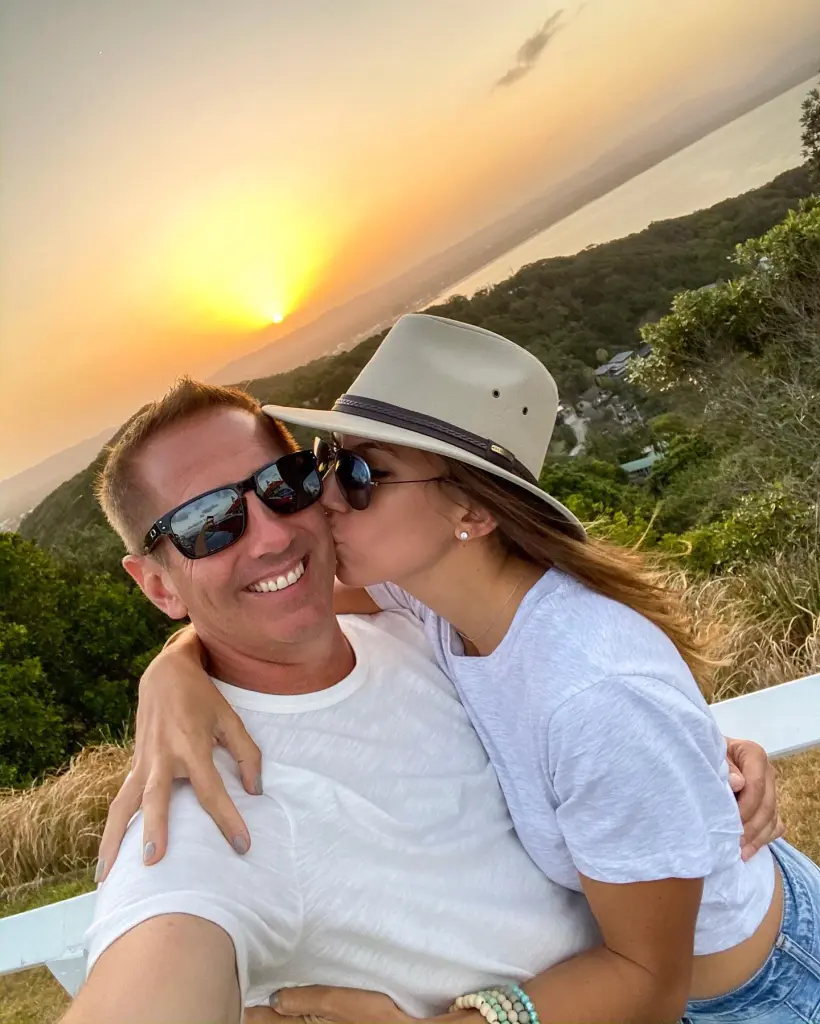 NASCAR driver Greg Biffle poses with his wife, who is kissing him, with a sunset and scenic background.