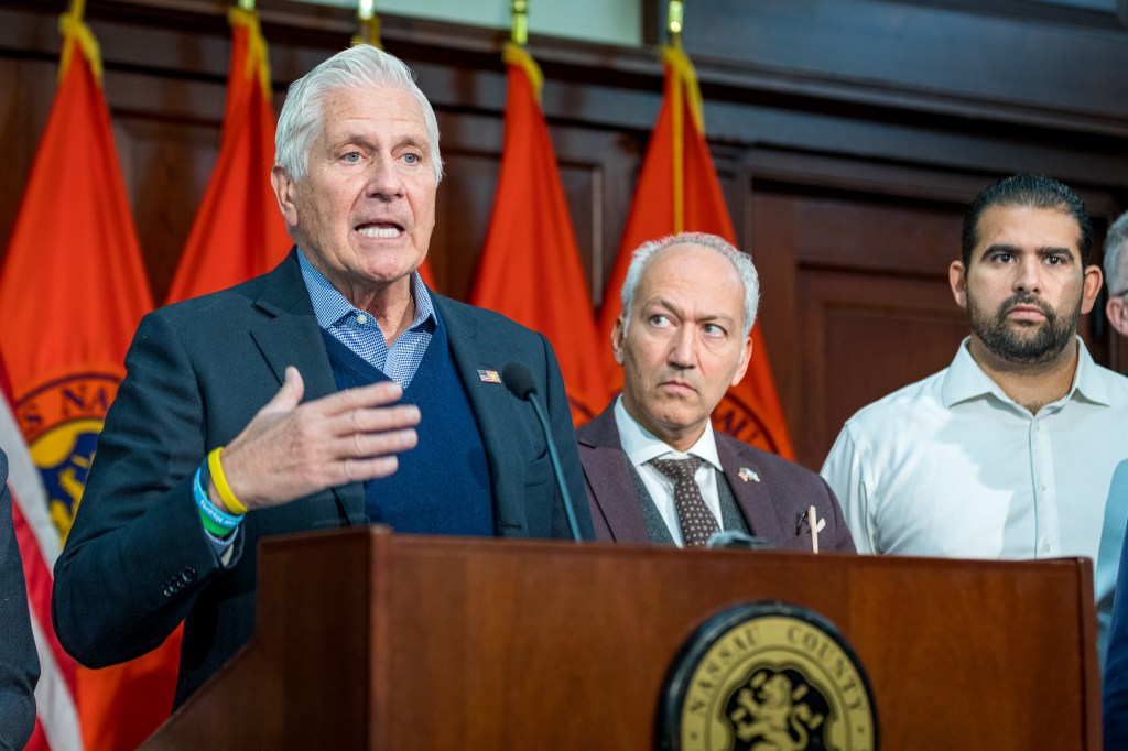 Nassau County Executive Bruce Blakeman speaks at a podium with two men standing behind him and red flags in the background.