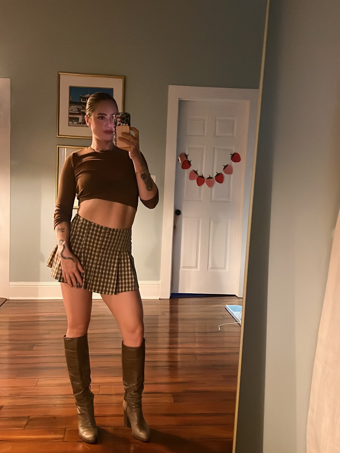 Natalie Tippett takes a mirror selfie in a brown crop top, plaid skirt, and knee-high boots.