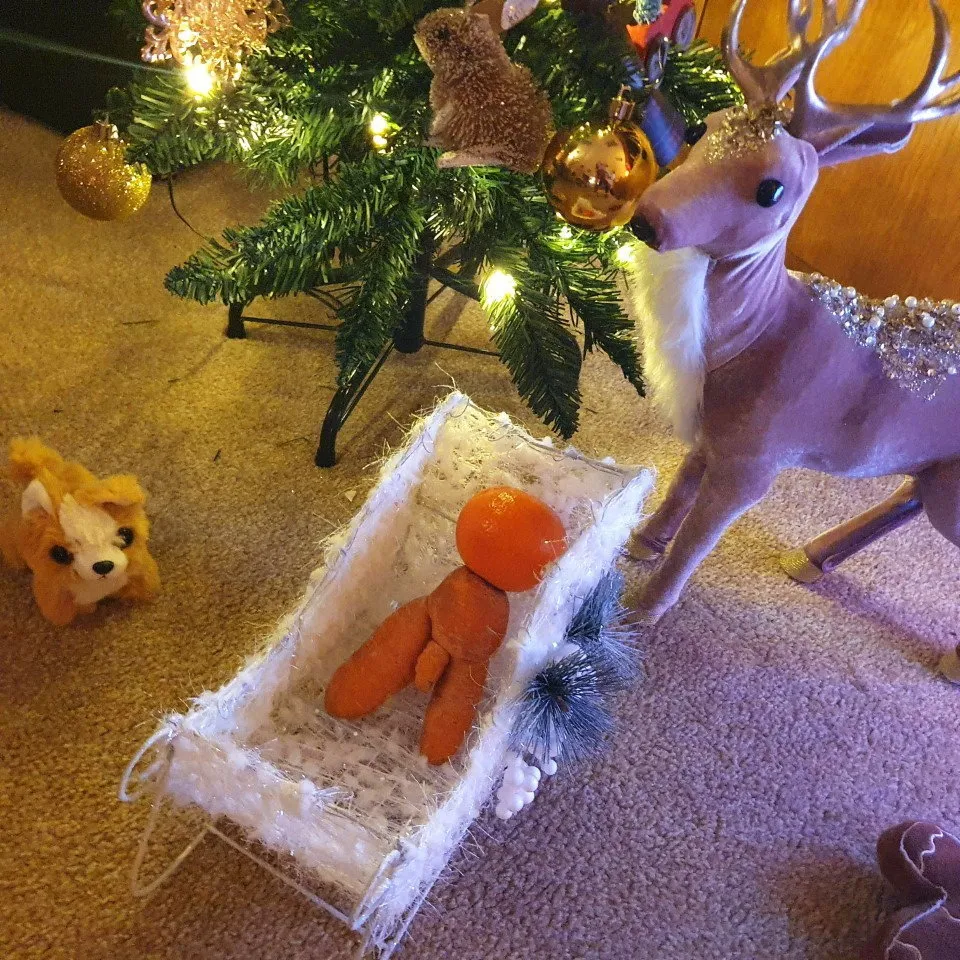 A naughty carrot with a tangerine head in a white sleigh by a Christmas tree.