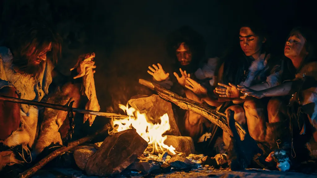 Family of prehistoric hunter-gatherers cooking meat over a bonfire in a dark cave.