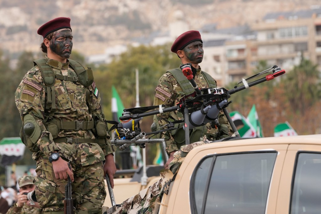 Two New Syrian Army soldiers, wearing camouflage and maroon berets, with painted faces, stand next to an armored drone during a parade.