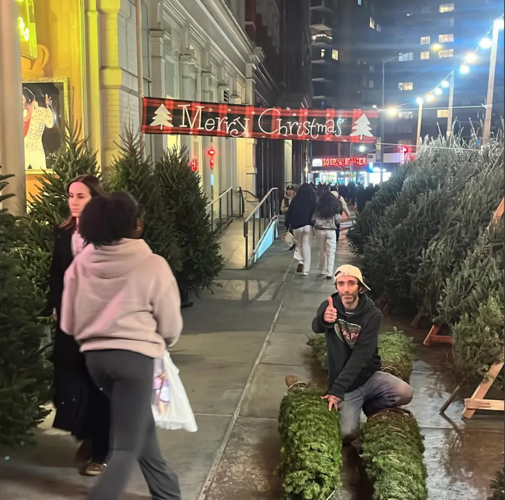 A man kneels on a New York City sidewalk in front of two bundled Christmas trees, giving a thumbs-up.