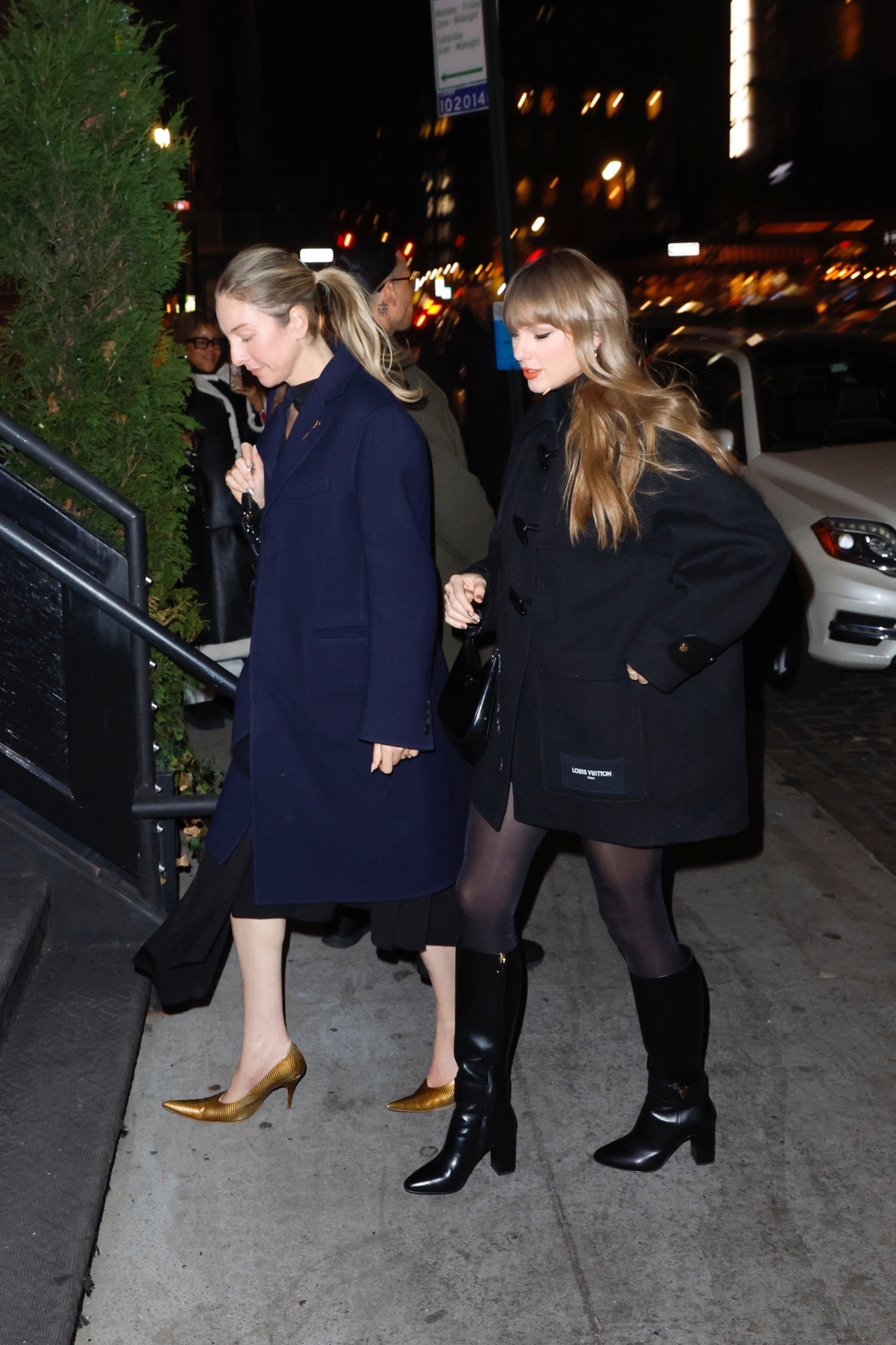 Taylor Swift in a black coat and boots with a friend in a navy coat and gold heels, stepping out of a restaurant at night.