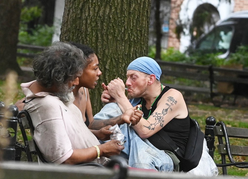 Three individuals, including a man with a blue headwrap, using drugs in a park.