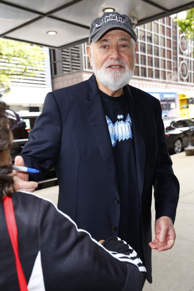 Rob Reiner wearing a Spinal Tap baseball cap and a black t-shirt under a black blazer while signing an autograph for someone holding a blue marker.