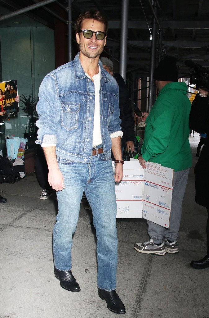 Glen Powell wearing a denim jacket and jeans, smiling while exiting CBS Mornings in NYC.