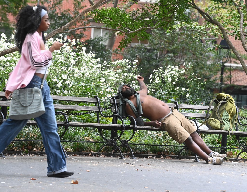 A homeless man sleeping on a park bench as a woman walks past.