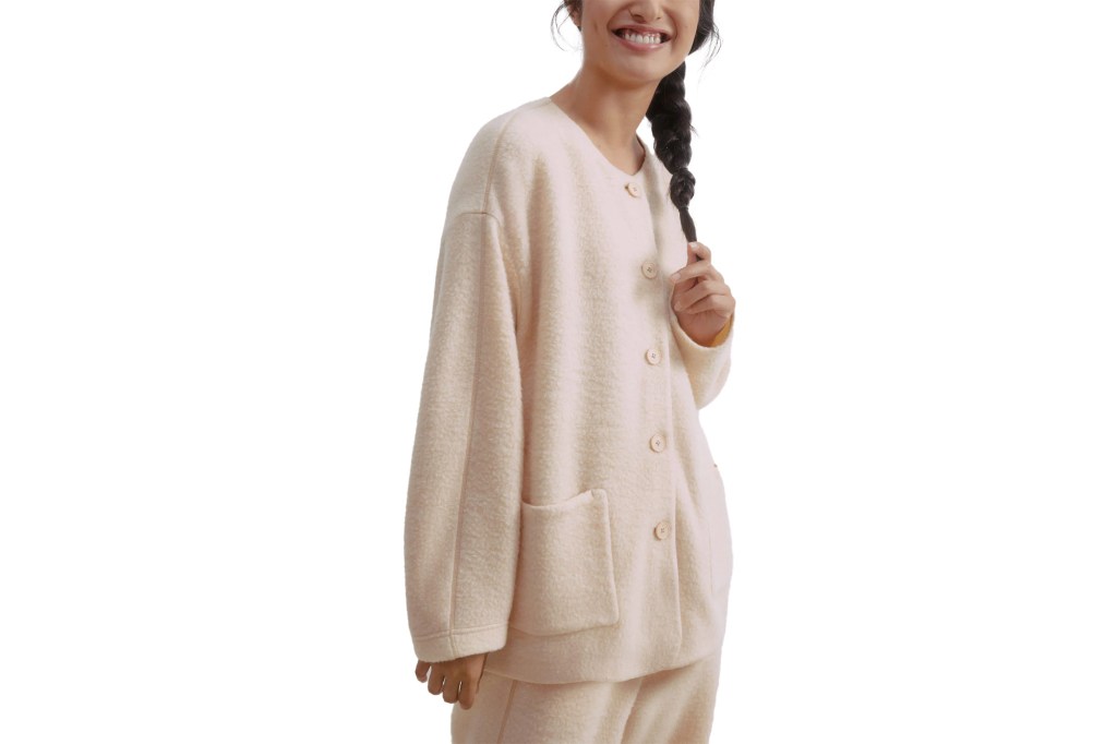 A woman wearing a cozy button-up cream colored jacket and matching pants, smiling as she holds her braided hair.