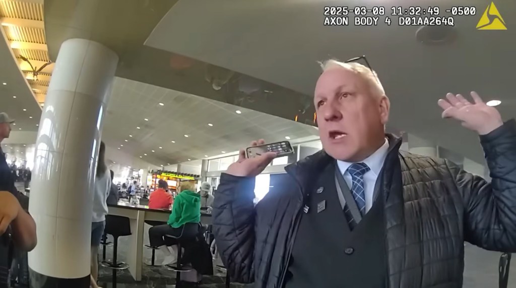 Body camera footage shows a male flight attendant holding a phone while speaking with his hands raised, with other people and airport architecture visible in the background.