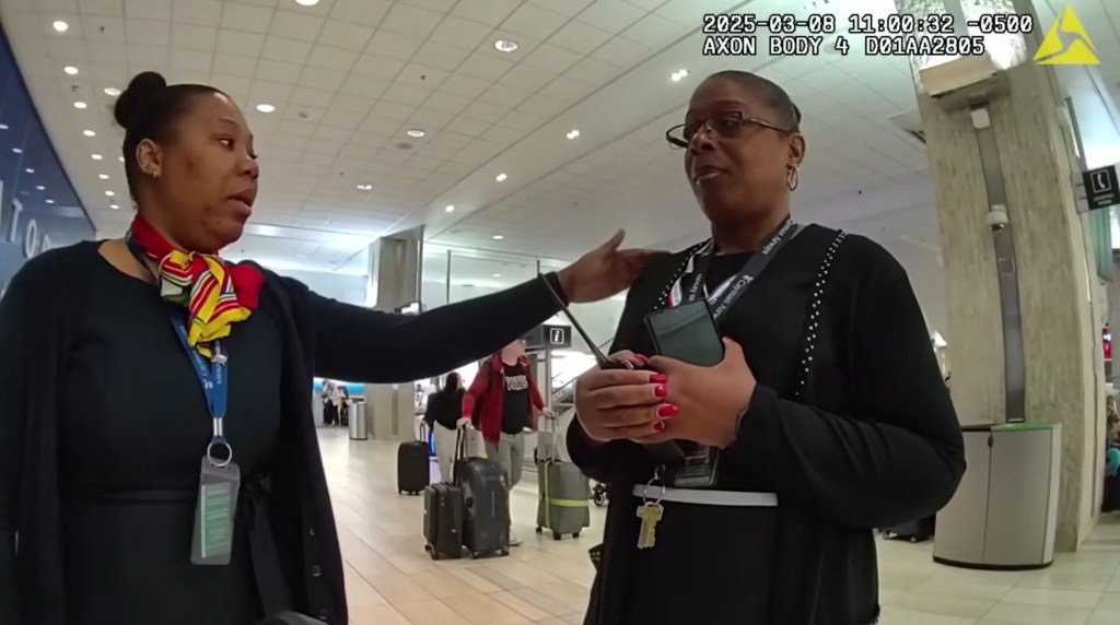 Two airline employees, wearing uniforms and lanyards, are conversing in an airport terminal.