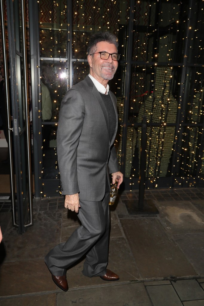 Simon Cowell wearing a gray suit, white shirt, and dark sweater, holding a bottle.